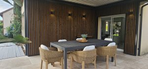 Varanda com revestimento ripado vertical WoodUpp em madeira escura com moldura preta, iluminação embutida, e piso de deck WoodUpp cinza-escuro à esquerda. Mesa de jantar cinza e cadeiras de vime complementam o espaço, criando área externa elegante e acolhedora e moderna.