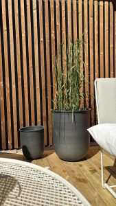 Painel ripado vertical e piso de madeira WoodUpp: ripas de madeira tratada em tom acobreado formando um revestimento elegante, sobre deck de tábuas WoodUpp. Em primeiro plano, dois vasos cinza com relva alta e uma cadeira clara, sob luz solar direta.