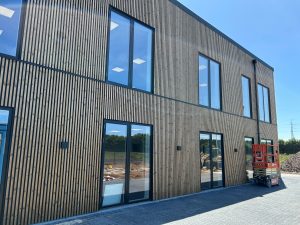 Fachada de dois andares com grandes vãos envidraçados e revestimento WoodUpp composto por ripas de madeira verticais estreitas em acabamento natural, espaçadas uniformemente e montadas em painéis, com perfis metálicos escuros. No chão, plataforma elevatória vermelha e piso de blocos intertravados.