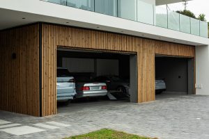 Fachada de casa moderna com sacada de vidro, três portas de garagem abertas e carros no interior; revestimento ripado de madeira WoodUpp cobrindo pilares e laterais das garagens, destacando painéis verticais WoodUpp como acabamento elegante sobre entrada com piso de pedras e pequeno gramado.