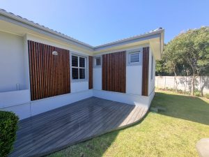 Fachada branca com painéis verticais de ripas de madeira WoodUpp em tom castanho e um deck WoodUpp cinza com canto arredondado; janela e luminária externa, relvado e árvores ao fundo, mostrando uma instalação exterior elegante e contemporânea dos produtos WoodUpp.