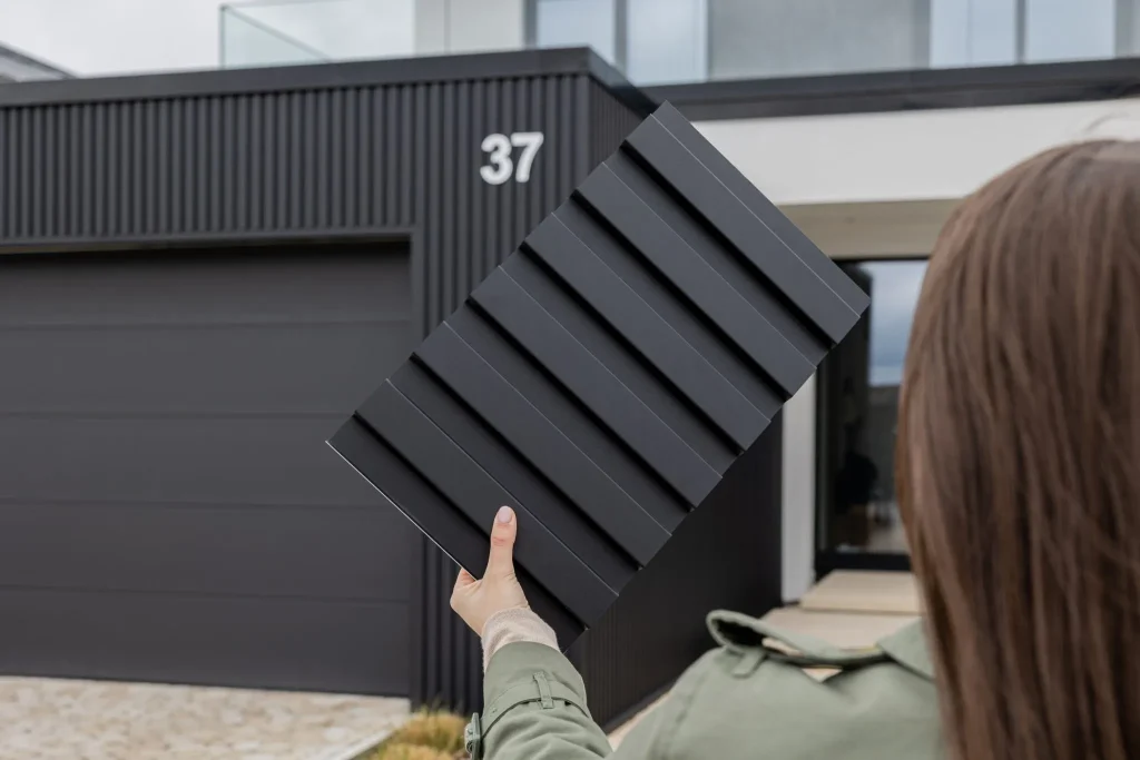Pessoa segurando uma amostra de painel ripado antracite da WoodUpp em frente a uma casa moderna com revestimento externo WoodUpp semelhante e porta de garagem antracite. O painel mostra perfis horizontais profundos, acabamento fosco e textura uniforme, ideal para fachadas contemporâneas.