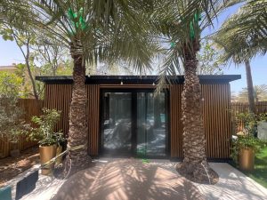 Cabana de jardim WoodUpp com revestimento ripado em madeira natural, painéis modulares WoodUpp e caixilho preto, portas de vidro de correr. Dois coqueiros em primeiro plano, vasos com plantas e luzes pendentes, sobre pavimento claro e sombras das palmeiras compõem o ambiente exterior ensolarado.