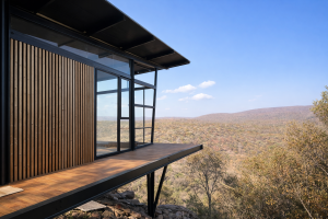 Uma vista exterior de uma cabana moderna elevada. A fachada apresenta os painéis ripados de madeira da WoodUpp, com ripas verticais de tom quente a cobrir a parede ao lado de grandes janelas com caixilharia preta e um deck de madeira, sobre uma vasta paisagem natural.