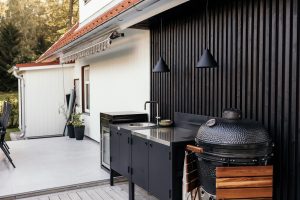 Área externa com cozinha WoodUpp: módulo preto com bancada inox e pia embutida, frigobar integrado, luminárias pendentes pretas e churrasqueira tipo kamado WoodUpp com tábuas laterais em madeira. Parede de ripas pretas ao fundo e piso em deck e cerâmica clara.