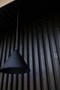 Detalhe de parede ripada em madeira escurecida WoodUpp, com painéis verticais e forro ripado no teto; superfície com nós e veios visíveis. À frente, pendente cônico preto desfocado. Produto WoodUpp: revestimento ripado vertical e forro ripado em madeira tratada com acabamento natural.