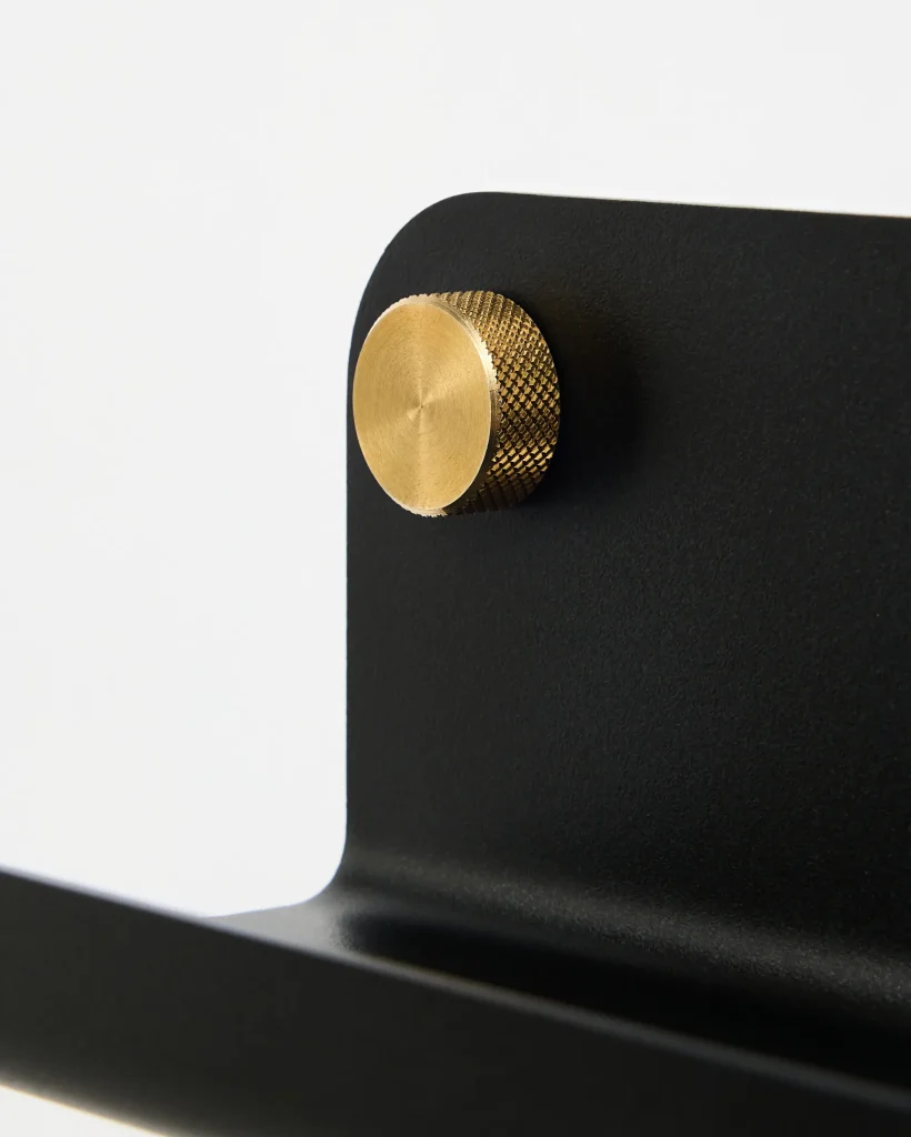 Detalhe de produto WoodUpp: suporte em metal preto fosco com canto arredondado e acabamento curvo, acompanhado de um parafuso/botão canelado em latão dourado texturizado que fixa as peças. Contraste elegante entre o preto opaco e o botão metálico brilhante visível