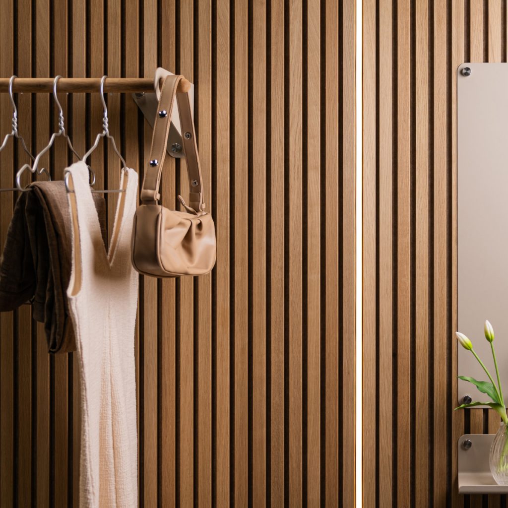 Painel ripado em madeira WoodUpp com lâminas verticais e textura natural ocupa a imagem; varão/cabideiro WoodUpp com suportes metálicos sustenta vestidos claros, casaco escuro e bolsa bege; à direita, espelho e pequena prateleira metálica com vaso de flores completam o conjunto.