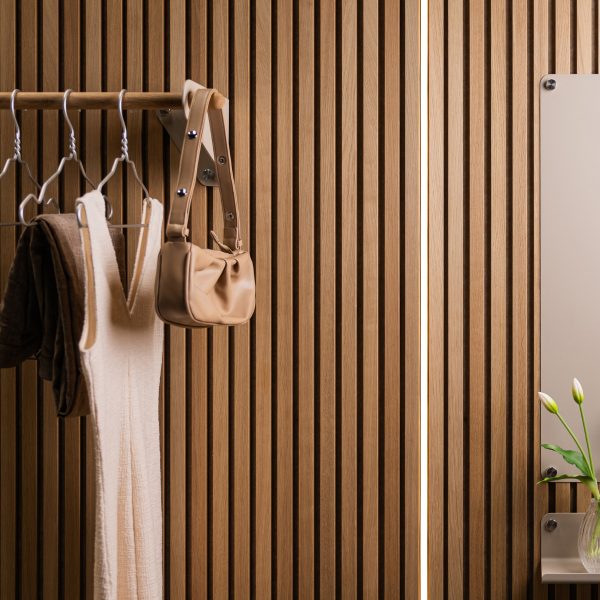 Painel ripado em madeira WoodUpp com lâminas verticais e textura natural ocupa a imagem; varão/cabideiro WoodUpp com suportes metálicos sustenta vestidos claros, casaco escuro e bolsa bege; à direita, espelho e pequena prateleira metálica com vaso de flores completam o conjunto.