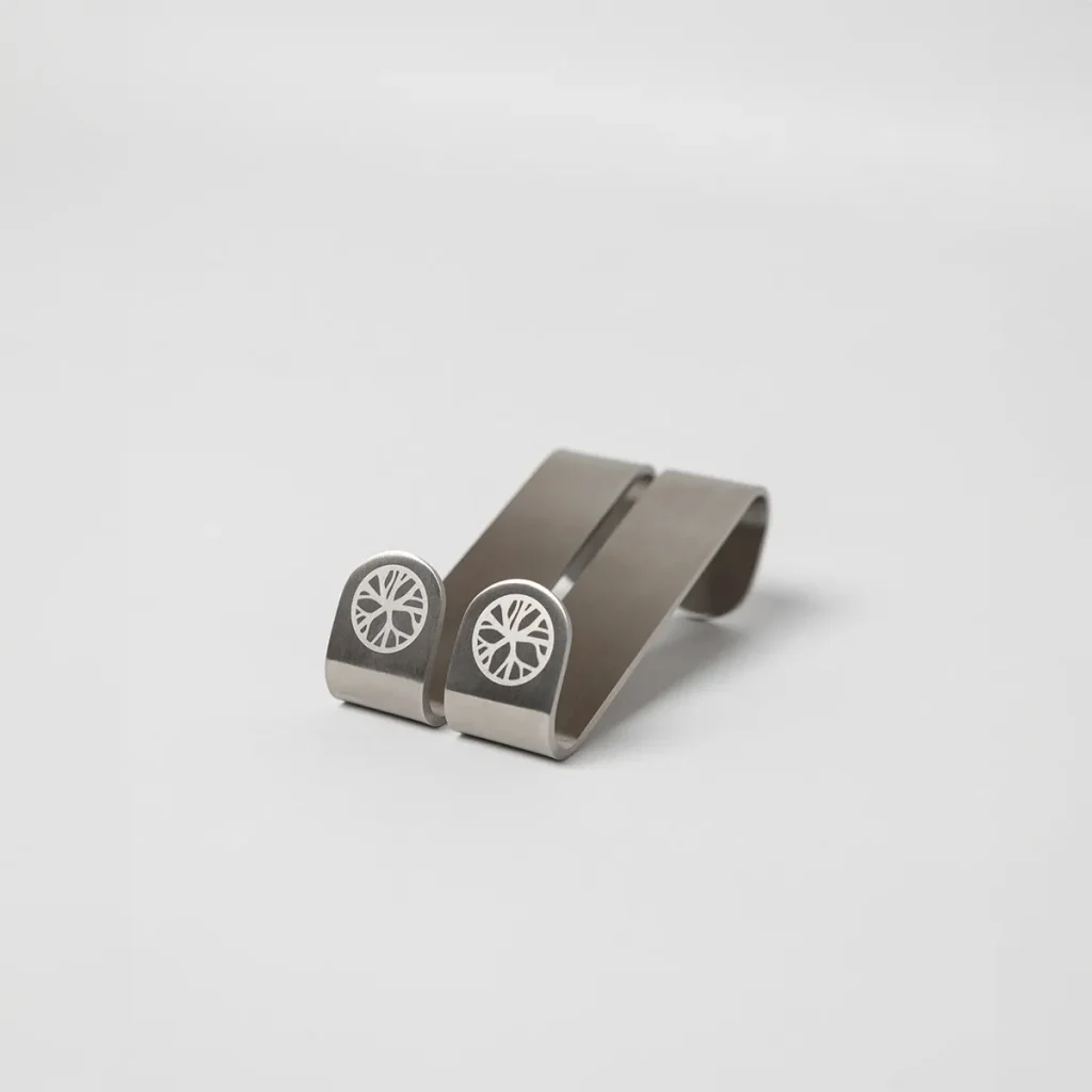 Imagem mostra dois clipes metálicos WoodUpp em aço escovado, de formato curvo e acabamento prateado, com gravura circular branca semelhante a uma roda na face frontal. Clipes compactos e minimalistas, projetados para prender objetos finos, exibidos sobre fundo branco neutro.