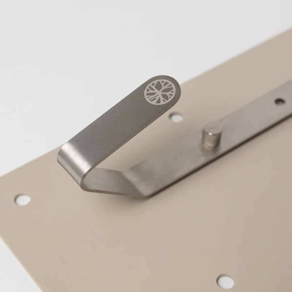 Imagem mostra um gancho de parede WoodUpp em aço inox escovado, com design curvado e logotipo gravado circular na ponta. O gancho está preso a uma placa perfurada bege por um parafuso de ajuste visível, evidenciando acabamento minimalista e aparafusamento robusto.