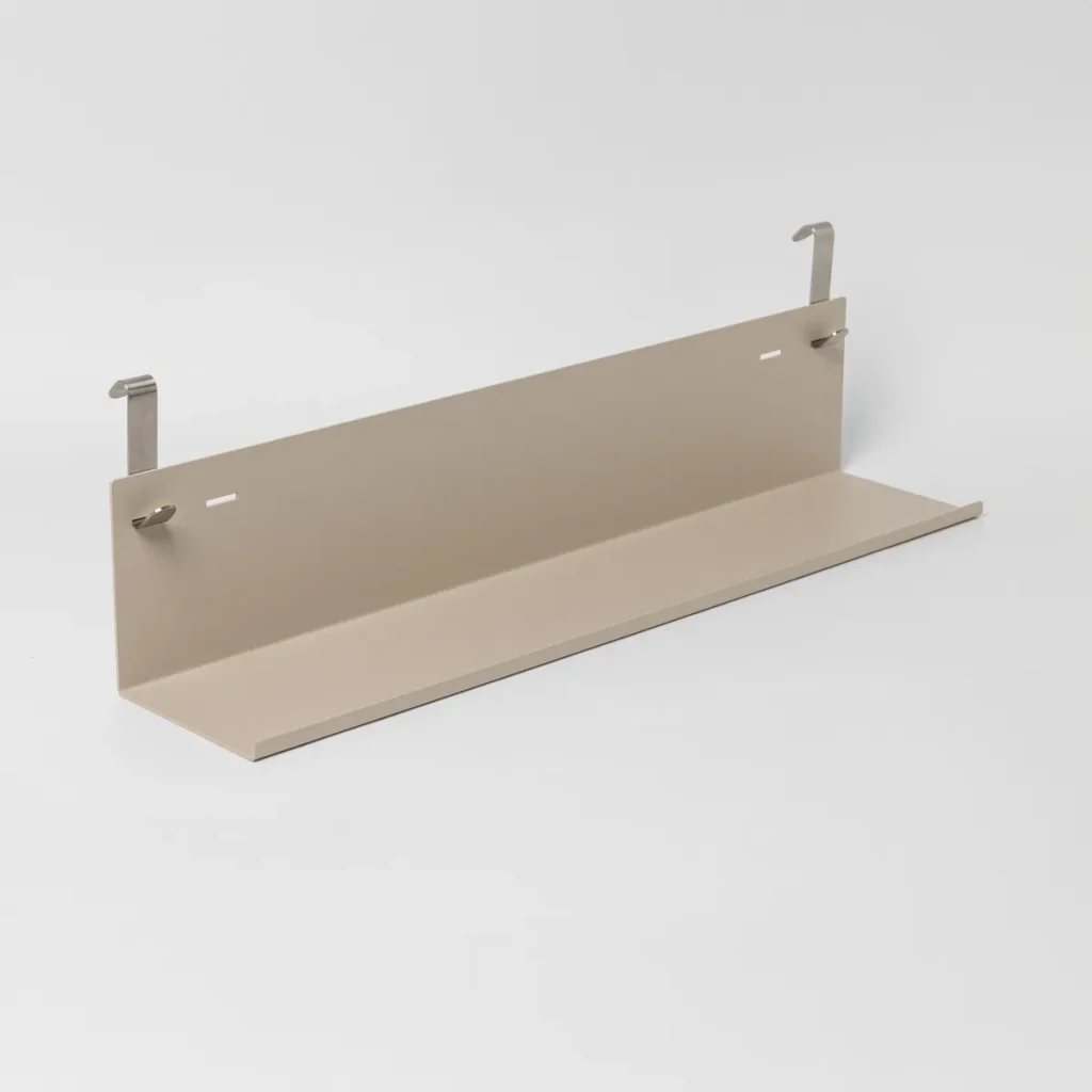 Prateleira metálica bege da WoodUpp em formato de L, com borda frontal elevada para apoio de objetos, acompanhada por dois ganchos metálicos prateados para pendurar em painéis. Superfície lisa e acabamento fosco, design minimalista, compacta e resistente, com fixação simples, ideal para organização de pequenos objetos.