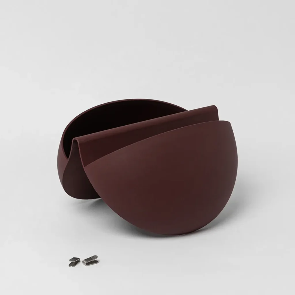 Imagem mostra produto WoodUpp: vaso de parede modular em duas peças curvas encaixadas, cor marrom escuro com acabamento fosco, formando um compartimento interno circular. Acompanha dois pequenos ganchos/peças metálicas para fixação. Design orgânico e minimalista, adequado para plantas ou decoração.