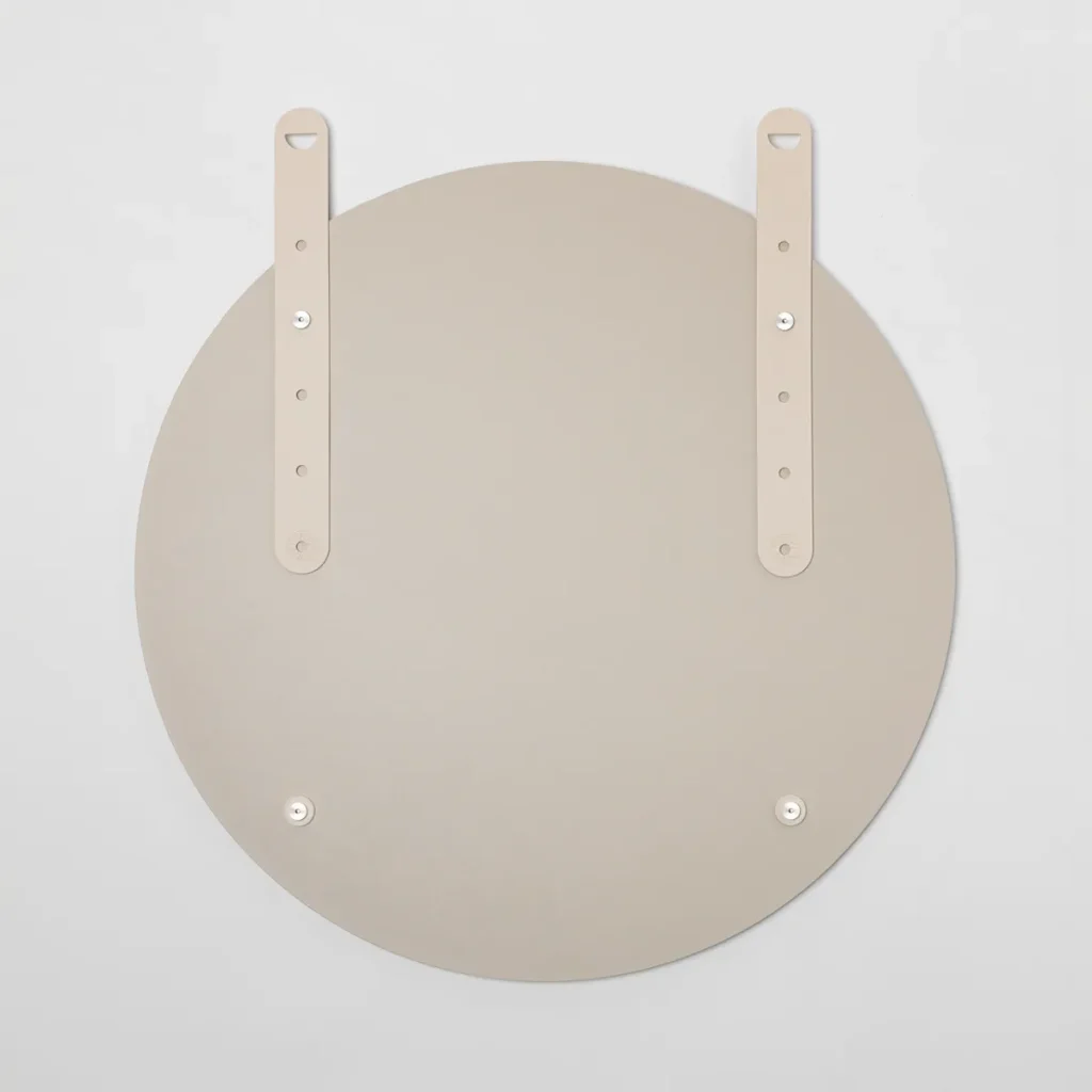 Parte traseira de um espelho redondo WoodUpp em tom bege claro, mostrando duas tiras de couro ajustáveis combinando, com fivelas e orifícios de regulagem, e quatro fixadores metálicos/discos magnéticos prateados visíveis na superfície, destacando o sistema de suspensão e acabamento minimalista do produto.