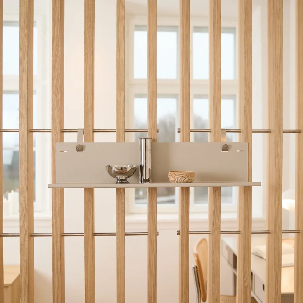 Divisória vertical de ripas de madeira da WoodUpp com trilhos metálicos, equipada com uma prateleira modular WoodUpp em acabamento neutro. Na prateleira, um vaso metálico, um cilindro prateado e uma tigela pequena de madeira completam a composição minimalista e funcional.
