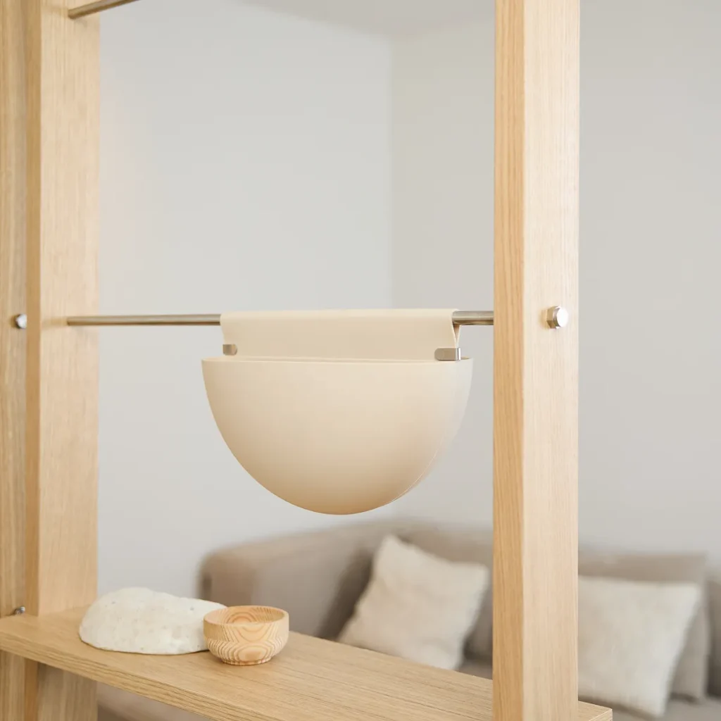 Estante modular WoodUpp em madeira clara com trilho metálico, exibindo um vaso pendente WoodUpp bege em forma de meia‑lua preso ao suporte. Na prateleira há pequenos objetos decorativos: tigela de madeira e pedra. Ambiente minimalista e acolhedor em tons neutros, acabamento escandinavo.