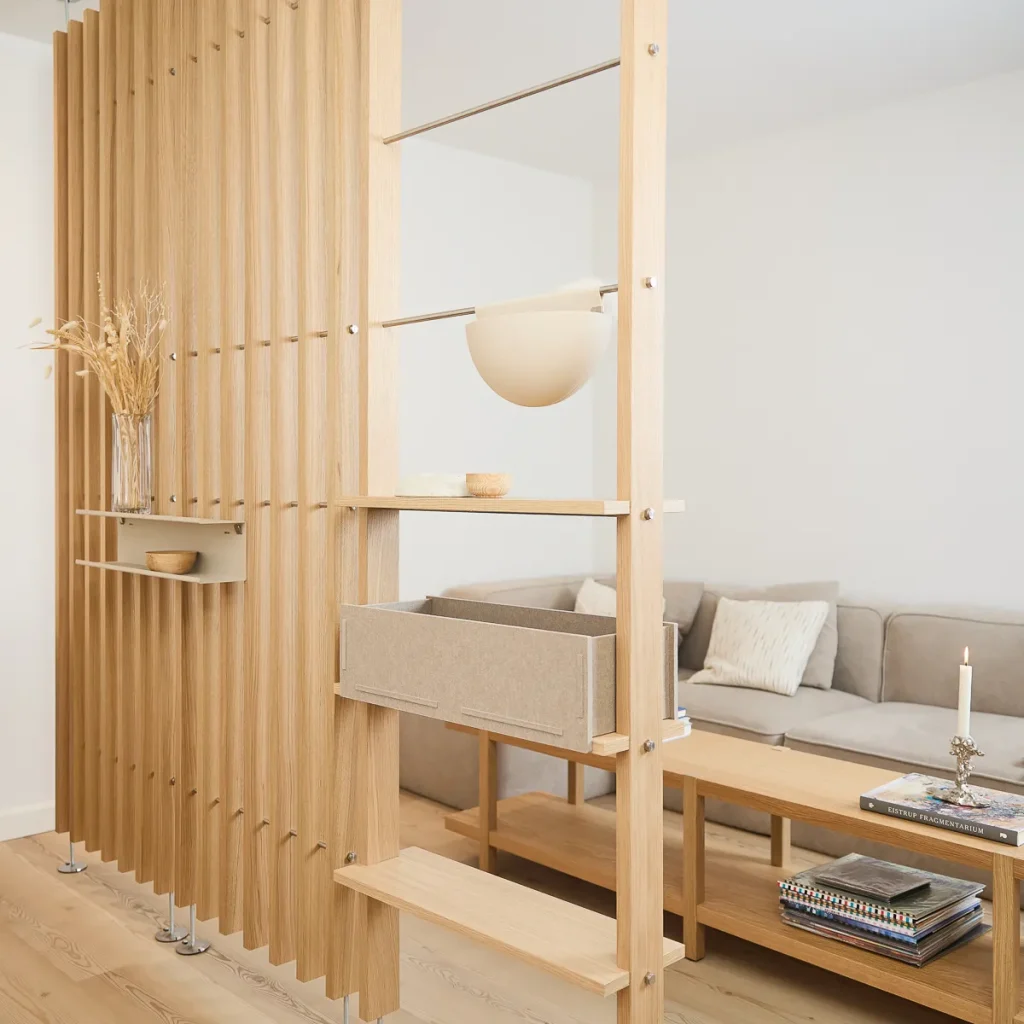 Divisória modular WoodUpp em lâminas de madeira clara com suportes metálicos; prateleiras ajustáveis, um baú/caixa de tecido suspenso, pequena prateleira lateral com vaso de flores secas e tigela. Ao fundo, sofá neutro e mesa de madeira com velas e livros.