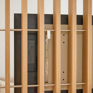 Painel ripado WoodUpp em carvalho natural com barras horizontais metálicas, mostrando a montagem traseira de uma TV presa ao painel pelo suporte de TV WoodUpp em aço escovado; detalhe do fecho e do perfil metálico entre ripas, vista próxima e minimalista