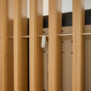 Painel WoodUpp com réguas verticais de madeira clara (carvalho) formando um brise interno, barra metálica horizontal e gancho de metal prateado encaixado, apoiado sobre backing perfurado bege. Mostra acabamento natural, encaixes modulares e acessório de pendurar da coleção WoodUpp, elegante.