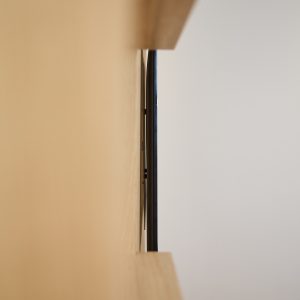 Imagem mostra painel claro da WoodUpp com duas prateleiras em madeira e uma televisão de borda fina montada rente ao painel; vê-se lateralmente o painel e o suporte da TV, evidenciando acabamento minimalista e encaixe estreito entre a tela e a madeira.