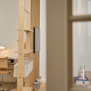 Divisória de madeira WoodUpp com prateleiras abertas, caixa de arrumação branca e suporte de TV embutido, organização com prateleira metálica inferior para controles; ao fundo, canto de escritório com cadeira de palhinha e mesa, ambiente minimalista e iluminado com livros e vela decorativa.