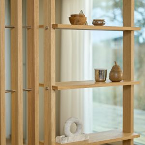 Estante modular WoodUpp em carvalho claro, com ripas verticais, prateleiras flutuantes e barras metálicas de fixação aparentes; exibe objetos decorativos: tigelas de madeira, um pequeno copo metálico, recipiente em forma de pera em madeira e uma escultura circular em mármore sobre a prateleira.