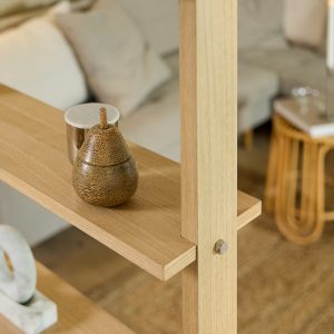 Imagem da estante modular WoodUpp em carvalho claro: estrutura vertical com prateleiras finas encaixadas por parafusos, de design escandinavo, acabamento natural e cantos retos. Na prateleira, objetos decorativos (pêra esculpida em madeira e vaso cerâmico), em ambiente de sala neutra e luminoso, aconchegante.