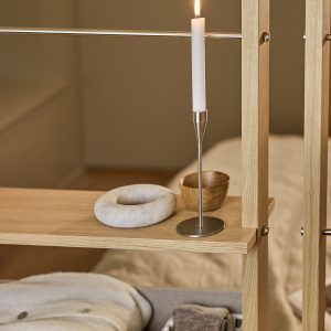 Estante divisória da WoodUpp em madeira clara com prateleiras e hastes metálicas; sobre uma prateleira aparecem um castiçal fino de aço inox com vela branca acesa, uma tigela pequena em madeira e um objeto decorativo em mármore em formato de rosca, tudo sobre acabamento natural.