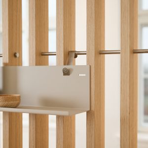 Divisória modular WoodUpp em ripas de carvalho claro com barra metálica horizontal, suportando uma prateleira metálica suspensa WoodUpp presa por um gancho/clip também metálico; pequena tigela de madeira sobre a prateleira. Acabamento minimalista e tons neutros, aparência funcional e elegante.