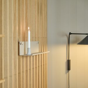 Prateleira de parede minimalista, compacta e discreta, e castiçal da WoodUpp em metal e cimento, montados num painel ripado de madeira clara, com vela branca fina acesa sobre a superfície. Ambiente com luminária de parede preta ao lado, criando iluminação suave.