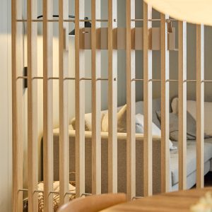 Divisória WoodUpp com ripas verticais de madeira clara unidas por hastes metálicas, separando uma sala com sofá cinza, almofadas e luminária. O produto WoodUpp mostra acabamento natural, espaçamento regular entre ripas e design modular elegante que preserva luminosidade e sensação de amplitude.