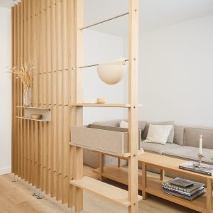 Painel divisório WoodUpp de ripas de madeira clara com prateleiras modulares e varões metálicos, exibindo uma caixa organizadora de tecido bege encaixada, pequenas prateleiras flutuantes e nicho para objetos; e um vaso com flores secas, ao fundo sofá e mesa de centro com livros e vela.
