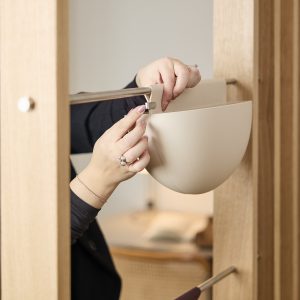 Mãos ajustando um vaso suspenso WoodUpp bege, preso por um gancho/clip de plástico ao suporte metálico horizontal fixado em estrutura de madeira clara. Mostra o vaso modular de formato semiesférico, o gancho de encaixe e o trilho metálico usados como sistema de organização.