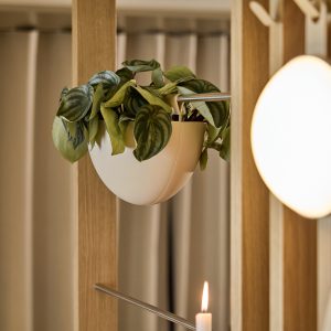 Vaso suspenso branco da WoodUpp preso a haste metálica do módulo divisório em ripas de madeira WoodUpp, com planta de folhas verdes variegadas caindo sobre a borda. À direita, luminária redonda e vela acesa completam a cena em ambiente minimalista.