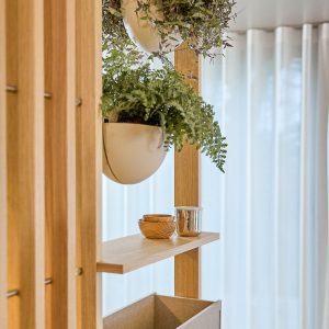 Imagem mostrando produtos WoodUpp: uma divisória modular em madeira clara com ripas verticais, prateleira e caixa de arrumação inferior, decorada com dois vasos suspensos brancos com folhagens, tigelas de madeira e um copo metálico sobre a prateleira, em fundo com cortinas translúcidas.