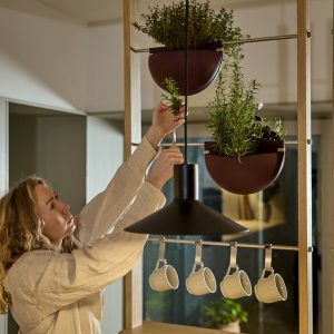 Mulher ajustando ervas em uma estante modular WoodUpp de madeira clara com varões metálicos; dois vasos suspensos meia‑lua bordô da WoodUpp cheios de plantas aromáticas, ganchos metálicos segurando quatro canecas brancas penduradas sob uma luminária preta, criando divisão funcional e estética na cozinha.