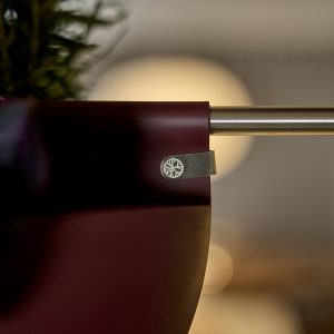 Close de um vaso suspenso WoodUpp vinho preso a uma barra metálica por um gancho/clip de metal prateado com logotipo gravado. Vaso curvo mostra planta desfocada no canto; clip WoodUpp segura firmemente o suporte, destacando acabamento e detalhe do emblema circular.