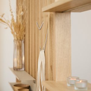 Painel ripado de madeira da WoodUpp em carvalho claro, com prateleiras flutuantes e ganchos metálicos prateados (modelo em V) fixados nas ranhuras; a imagem mostra uma tira de tecido pendurada num gancho, vela em porta-vidro e vaso com flores secas sobre as prateleiras.