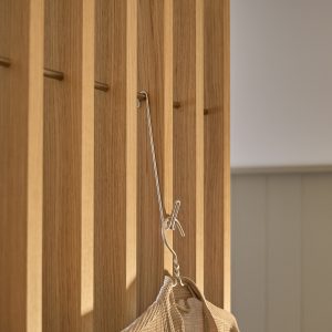 Painel ripado WoodUpp em carvalho claro com ripas verticais regulares; pequenos ganchos metálicos bronze inseridos entre as ripas; um cabide de arame prateado pendurado num gancho segura uma sacola/roupão de tecido bege, com acabamento liso e design minimalista, ideal para entradas.