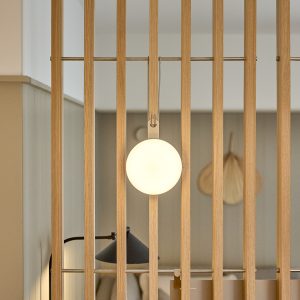 Divisória de ambiente WoodUpp composta por ripas verticais de madeira clara (carvalho) com acabamento natural e barra metálica dourada, e luminária WoodUpp de formato circular pendurada por alça metálica, difusor opalino que emite luz suave, valorizando o ambiente minimalista entre as ripas.
