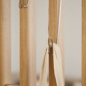Imagem detalhada de um elemento WoodUpp: ripado vertical em madeira clara com ganchos metálicos prateados WoodUpp fixados, usados como cabide. Uma alça de tecido bege está pendurada em um dos ganchos, mostrando acabamento minimalista e funcionalidade do mobiliário da marca.