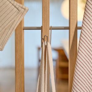 Imagem mostra produto WoodUpp: divisória de ripas em carvalho claro com barra metálica horizontal e dois ganchos em aço inox, usados para pendurar um pano de prato listrado, um pegador térmico bege e alças têxteis claras, em ambiente doméstico suave.