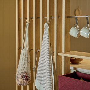 Painel ripado WoodUpp em madeira clara (carvalho) com varões metálicos e ganchos para pendurar, mostrando sacola de rede, panos de cozinha e canecas; inclui prateleira de madeira e caixa organizadora de feltro bordô da WoodUpp, em cena de cozinha funcional e elegante.