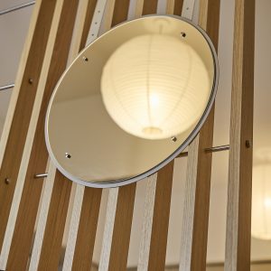 Painel ripado da WoodUpp em madeira clara com lâminas verticais alternadas, acompanhado de um espelho redondo WoodUpp preso por suportes metálicos e haste transversal. No espelho reflete uma luminária pendente de papel, destacando acabamento natural e conexão modular dos produtos WoodUpp.