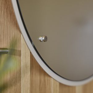 Close-up de espelho redondo WoodUpp preso por um imã metálico cromado, sobre parede de ripas de madeira clara WoodUpp; borda fina branca do espelho visível e um suporte tubular metálico à esquerda. Mostra acabamento e fixação dos produtos WoodUpp destacados.