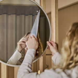 Pessoa remove película protetora de um espelho redondo WoodUpp, com moldura fina e acabamento neutro, montado sobre um painel de ripas de madeira claro WoodUpp; a imagem mostra as mãos e o reflexo da pessoa ao destacar a película, evidenciando o espelho e o revestimento em madeira.