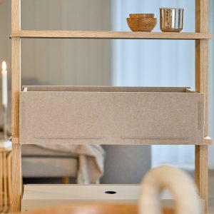 Estante modular de madeira clara WoodUpp com prateleiras minimalistas e uma gaveta/caixa de tecido bege WoodUpp encaixada no centro; no topo há duas tigelas de madeira e um copo metálico decorativo, em ambiente interno com luz suave e fundo neutro.