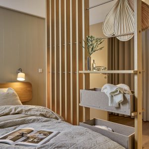 Quarto acolhedor com divisória de ripas e estante modular WoodUpp em madeira clara, com prateleiras, varões e caixas-gaveta de tecido WoodUpp; nas prateleiras vaso e objeto decorativo, nas caixas um suéter dobrado, ao lado cama com edredom neutro, abajur e revista aberta.