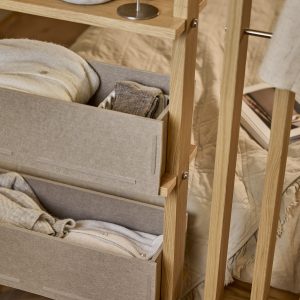 Imagem mostra um organizador vertical WoodUpp em madeira clara com duas caixas gaveta de feltro bege da WoodUpp, contendo roupas dobradas — suéteres e meias —, apoiado ao lado da cama com manta e revistas, prateleira superior com base metálica de abajur visível e tons neutros.