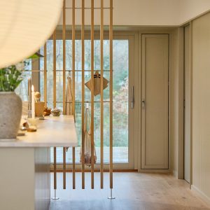 Divisória WoodUpp de ripas verticais em madeira clara, apoiadas no teto e no piso com bases metálicas, funcionando como cabideiro. Vêem-se ganchos WoodUpp com sacola de rede, panos e tábuas de corte pendurados, ao lado de bancada de cozinha com vasos e velas.